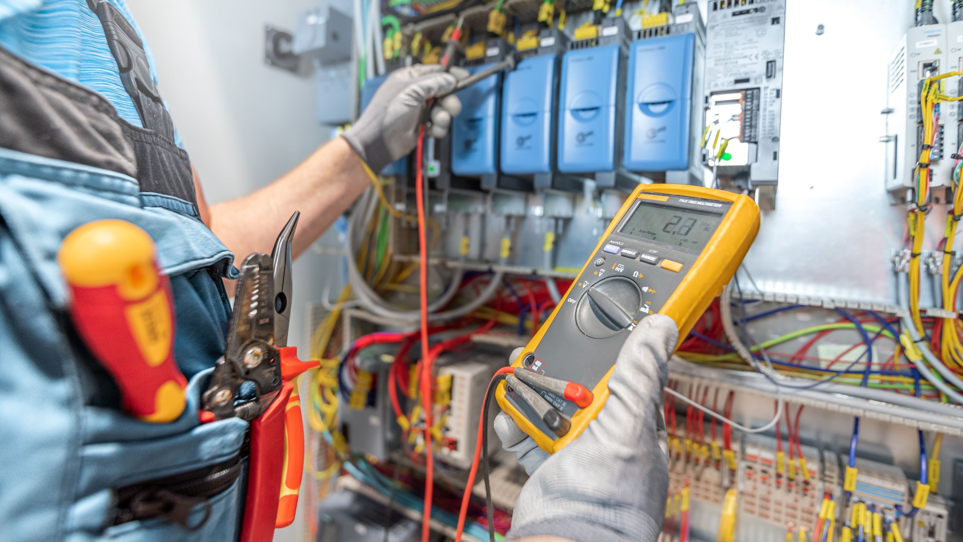 The electricians hand holding multimeter during checks the electrical wiring in the fuse box. Work in electrical industry.