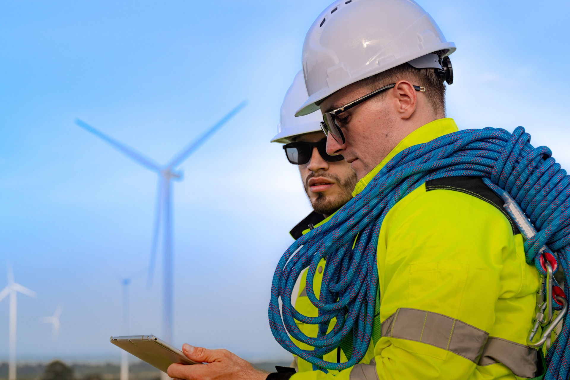 Workers in high visibility jackets examine a wind turbine farm, one pointing towards the turbines while the other holds a mobile device. The scene emphasizes renewable energy technological inspection.